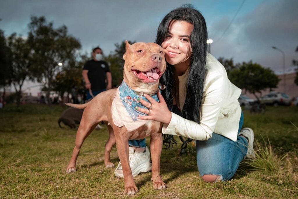 A woman with long hair smiles while hugging a pitbull in a Tacna park, enjoying a day outdoors.