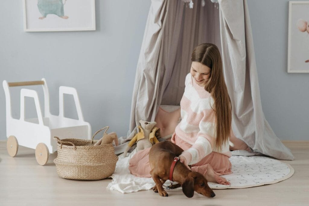 A smiling pregnant woman sits in a nursery, petting her dachshund dog.