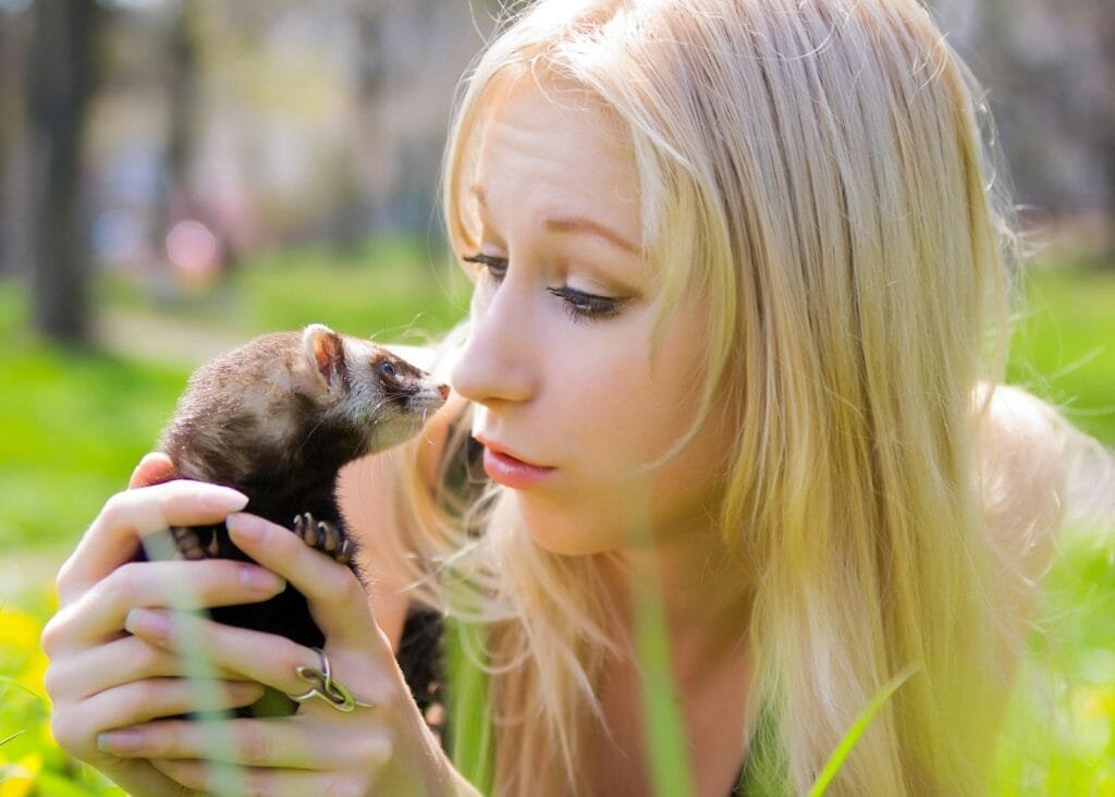 A young woman affectionately holding a ferret in a sunlit outdoor setting.