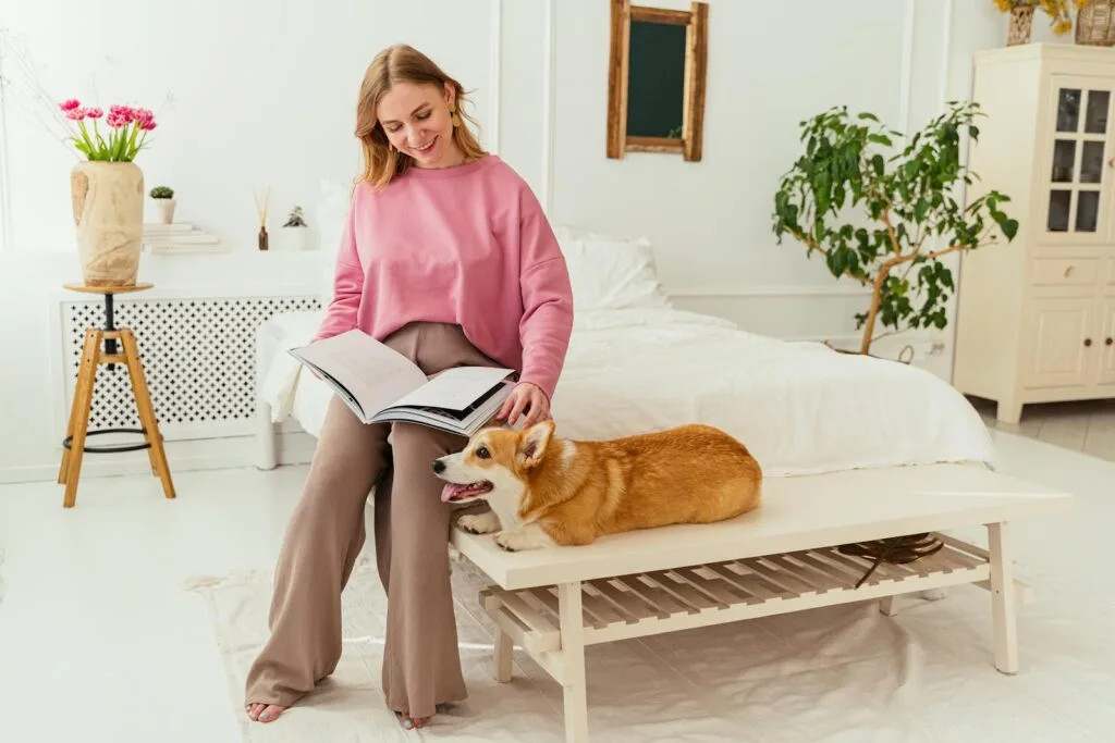 Woman in a pink sweater reads a magazine in a bright bedroom with a corgi on the bench.